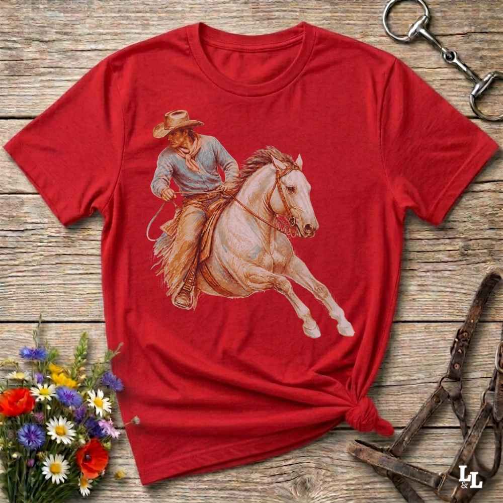 A red short-sleeve crewneck t-shirt featuring a western graphic of a cowboy riding a white horse, laid flat on a rustic wooden background with decorative flowers and tack.