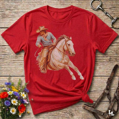 A red short-sleeve crewneck t-shirt featuring a western graphic of a cowboy riding a white horse, laid flat on a rustic wooden background with decorative flowers and tack.