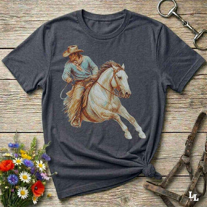 A dark heather gray t-shirt featuring a graphic print of a cowboy riding a white horse, styled in a flat-lay on a rustic wooden background with equestrian accessories and flowers.