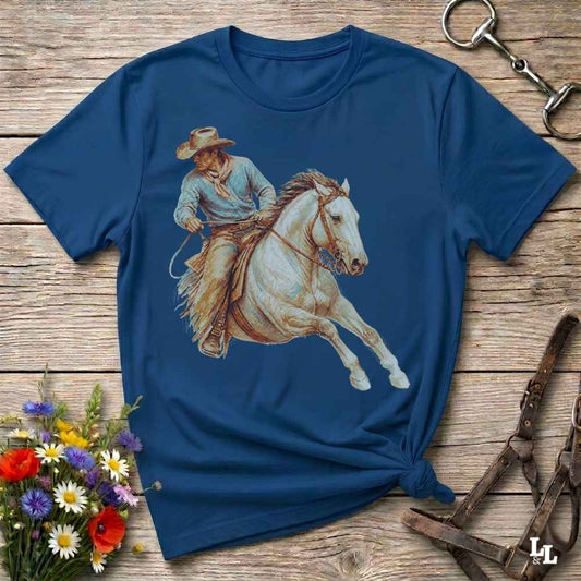 A navy blue short-sleeve t-shirt with a graphic print of a cowboy riding a white horse, laid flat on a rustic wooden surface with decorative flowers and horse tack.