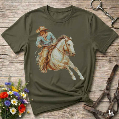 An olive green short-sleeve t-shirt with a graphic print of a cowboy riding a white horse, displayed on a rustic wooden background with decorative equestrian and floral props.