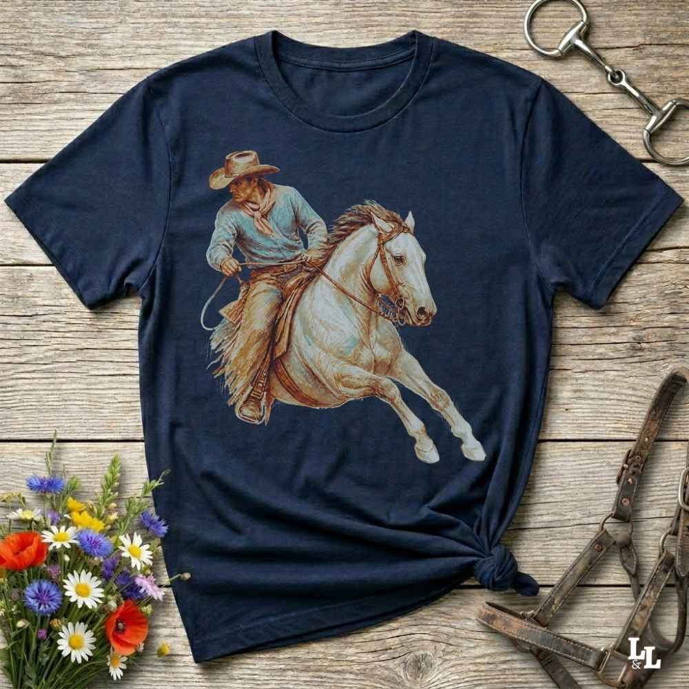 A navy blue short-sleeve t-shirt with a graphic print of a cowboy riding a white horse, styled on a rustic wooden background with equestrian accessories and flowers.