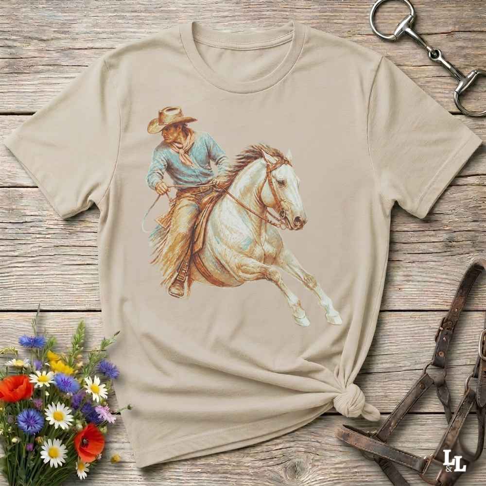 A beige short-sleeve t-shirt with a graphic print of a cowboy riding a white horse, styled in a flat-lay on a rustic wooden background with flowers and horse tack.