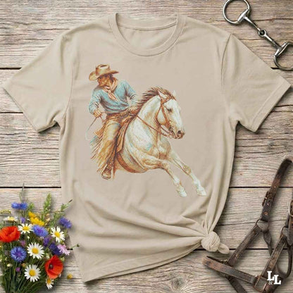 A beige short-sleeve t-shirt with a graphic print of a cowboy riding a white horse, styled in a flat-lay on a rustic wooden background with flowers and horse tack.