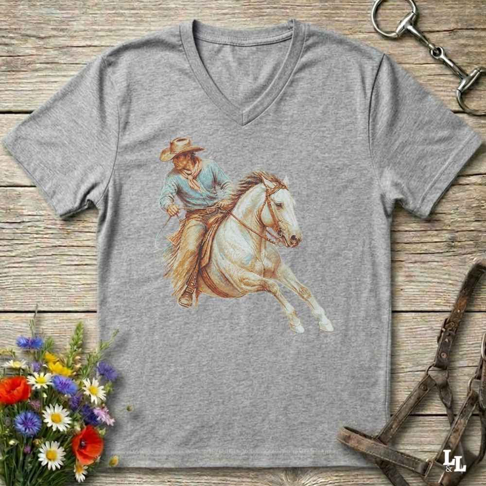 A heather gray V-neck t-shirt featuring a western-themed graphic of a cowboy riding a white horse, displayed on a rustic wooden background with decorative equestrian and floral props.