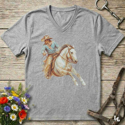 A heather gray V-neck t-shirt featuring a western-themed graphic of a cowboy riding a white horse, displayed on a rustic wooden background with decorative equestrian and floral props.