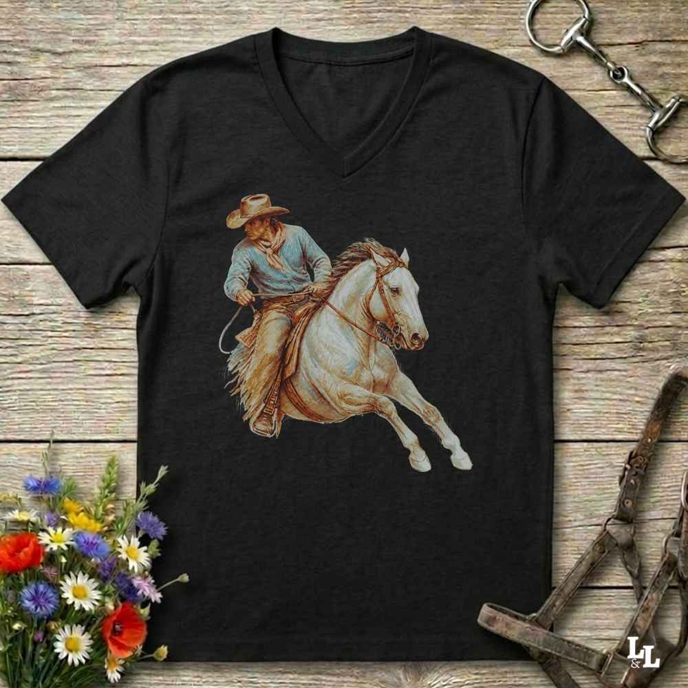 A black V-neck t-shirt featuring a graphic print of a cowboy riding a white horse, displayed on a rustic wooden background with equestrian and floral props.