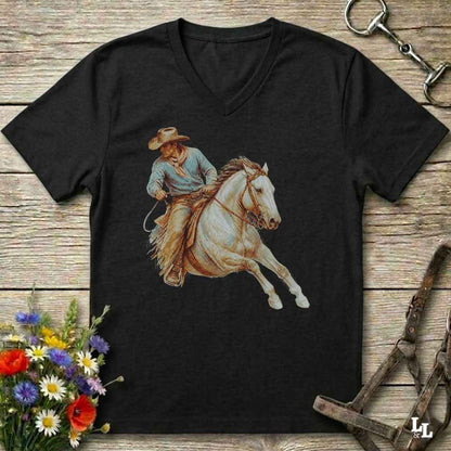 A black V-neck t-shirt featuring a graphic print of a cowboy riding a white horse, displayed on a rustic wooden background with equestrian and floral props.