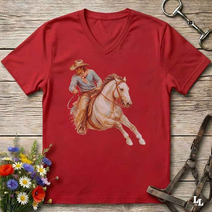 A red V-neck t-shirt featuring a western cowboy riding a white horse graphic, displayed flat on a rustic wooden background with equestrian and floral props.