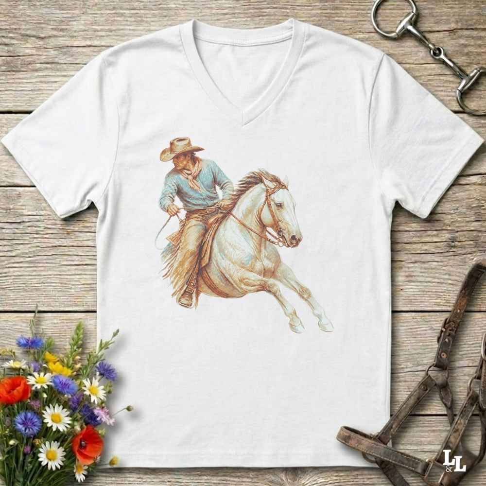 A white V-neck t-shirt laid flat on a rustic wooden surface, featuring a detailed graphic of a cowboy riding a white horse. The shirt is styled with equestrian accessories (a bit and reins) and a small bouquet of colorful wildflowers.