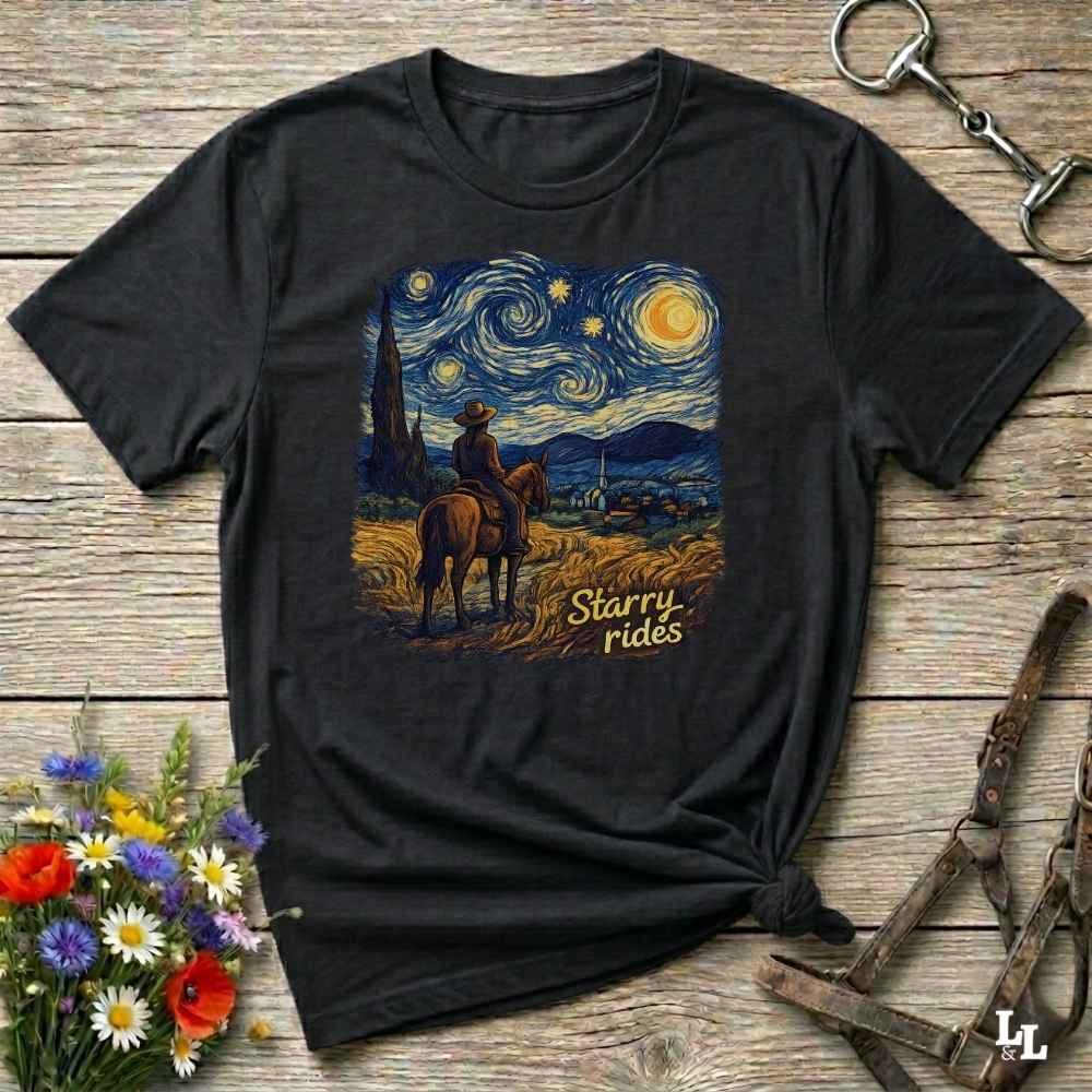 A black short-sleeve graphic T-shirt laid flat on a rustic wooden surface, styled with flowers, a horse bit, and leather tack. The shirt features a large central print of a cowboy on a horse under a swirling starry night sky, with the words 'Starry Rides' in yellow script.