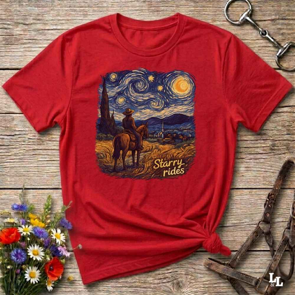 A red short-sleeve graphic T-shirt laid flat on a rustic wooden surface, styled with flowers, a horse bit, and a halter. The shirt features a large central print of a cowboy on a horse in a starry night landscape with the words 'Starry rides'.