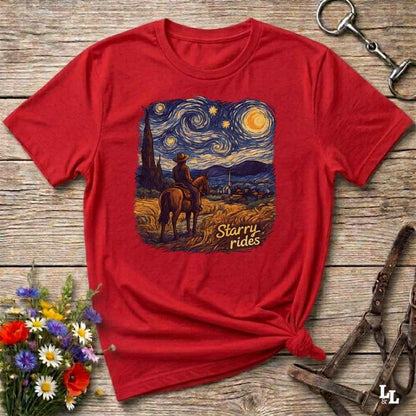 A red short-sleeve graphic T-shirt laid flat on a rustic wooden surface, styled with flowers, a horse bit, and a halter. The shirt features a large central print of a cowboy on a horse in a starry night landscape with the words 'Starry rides'.