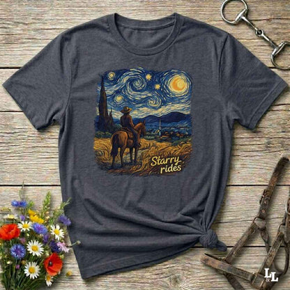 A dark heather gray graphic t-shirt laid flat on a rustic wooden surface, styled with a bouquet of flowers, a horse bit, and a leather halter. The t-shirt features a central graphic print in a Van Gogh-inspired style showing a cowboy on a horse under a swirling starry night sky, with the text 'Starry rides'.