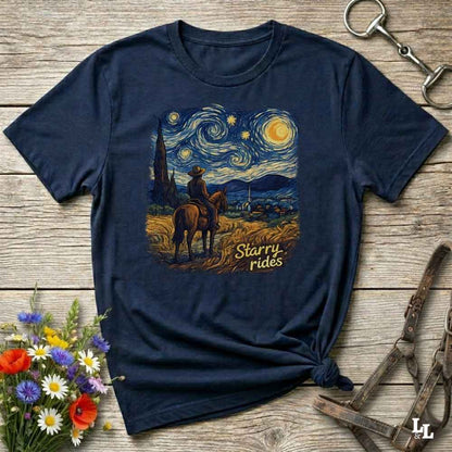 A navy blue graphic t-shirt laid flat on a rustic wooden surface, styled with a bouquet of wildflowers, a horse bit, and a leather bridle. The t-shirt features a central graphic print in a Starry Night-inspired style, showing a cowboy riding a horse through a field under a swirling blue and yellow sky, with the text 'Starry Rides'.