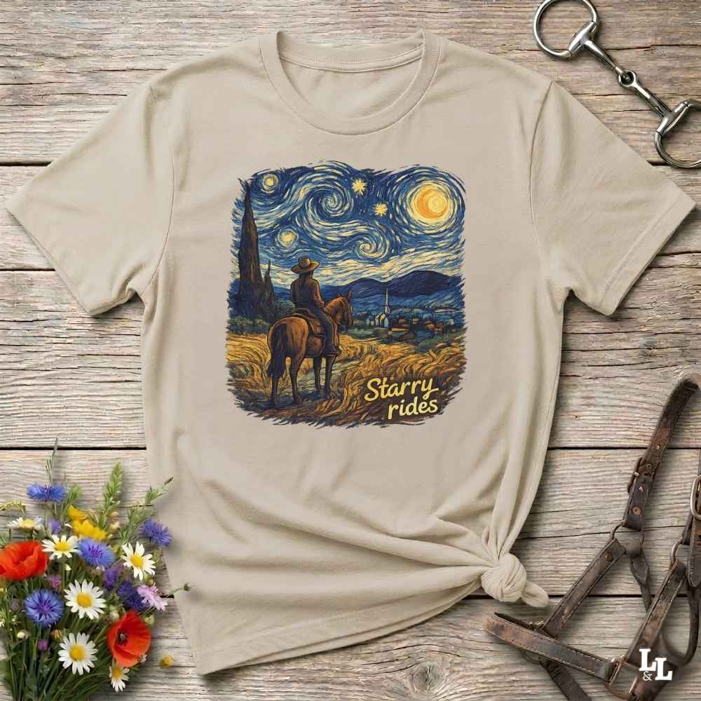 A beige graphic t-shirt styled in a flat-lay arrangement on a rustic wooden background, featuring a cowboy riding a horse under a swirling starry night sky with the text 'Starry rides'.