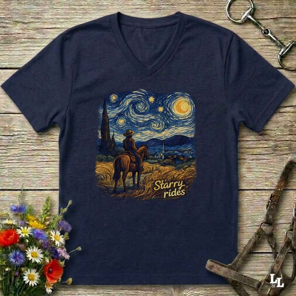 A navy blue V-neck t-shirt laid flat on a rustic wooden surface, featuring a graphic print of a cowboy riding a horse in a Starry Night style landscape with the text 'Starry rides'. The shirt is styled with equestrian tack in the background and a small bouquet of wildflowers in the lower left corner.
