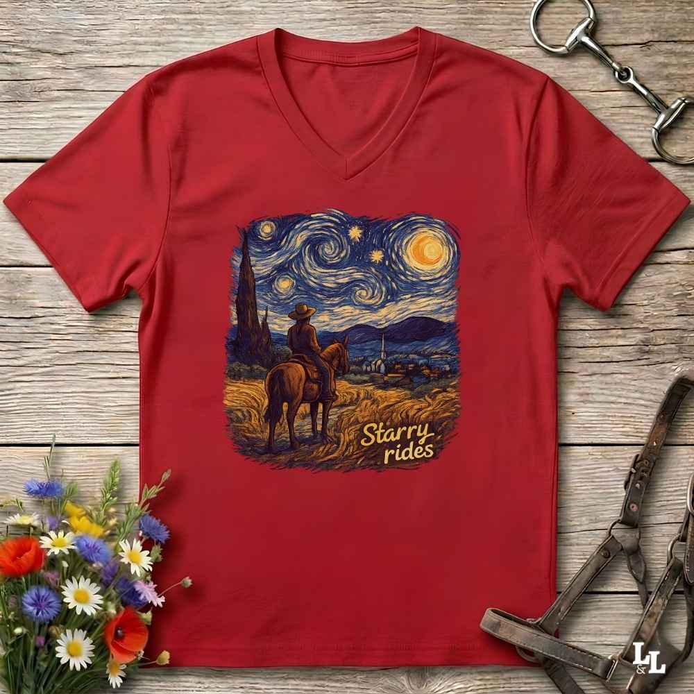 A red V-neck t-shirt laid flat on a rustic wooden surface, featuring a central graphic print of a cowboy riding a horse under a swirling Starry Night sky, with the text 'Starry rides'. The shirt is styled with equestrian tack in the top-right corner and a small bouquet of wildflowers in the bottom-left corner.