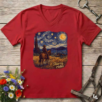 A red V-neck t-shirt laid flat on a rustic wooden surface, featuring a central graphic print of a cowboy riding a horse under a swirling Starry Night sky, with the text 'Starry rides'. The shirt is styled with equestrian tack in the top-right corner and a small bouquet of wildflowers in the bottom-left corner.
