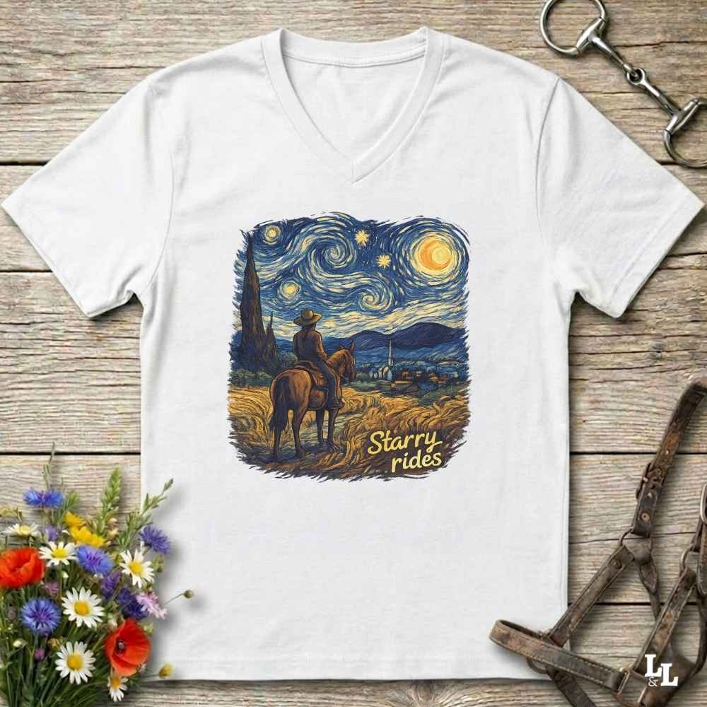 A white V-neck t-shirt laid flat on a rustic wooden surface, featuring a colorful graphic print of a cowboy riding a horse under a swirling starry night sky with the text 'Starry rides'. The shirt is styled with equestrian accessories (a leather bridle and a horse bit) and a small bouquet of wildflowers.