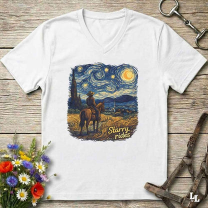 A white V-neck t-shirt laid flat on a rustic wooden surface, featuring a colorful graphic print of a cowboy riding a horse under a swirling starry night sky with the text 'Starry rides'. The shirt is styled with equestrian accessories (a leather bridle and a horse bit) and a small bouquet of wildflowers.