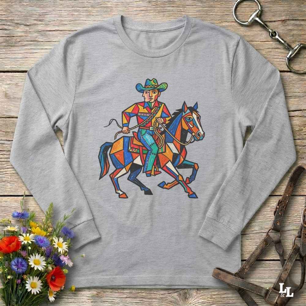 A light gray long-sleeve t-shirt with a colorful geometric cowboy on a horse graphic printed on the chest, displayed flat on a rustic wooden background with decorative flowers and leather tack.