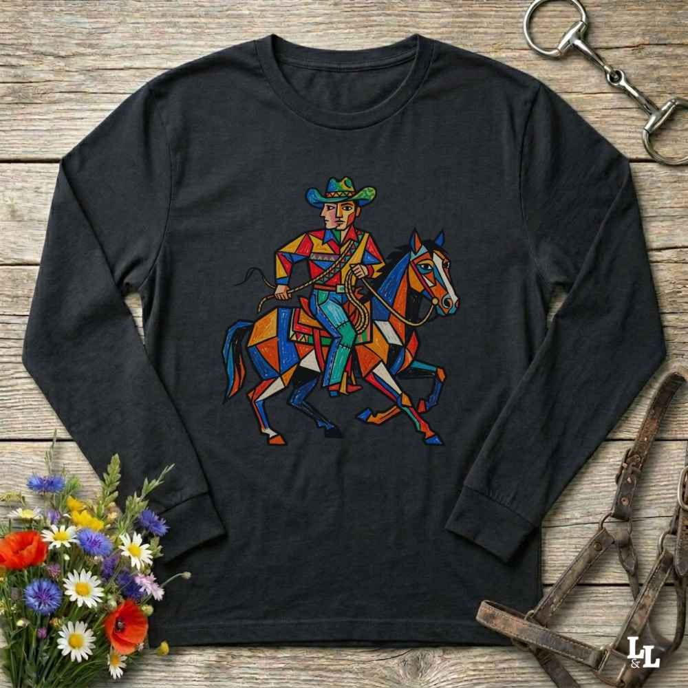 A black long-sleeve t-shirt with a colorful geometric graphic of a cowboy riding a horse, displayed flat on a rustic wooden background with flowers and leather tack props.