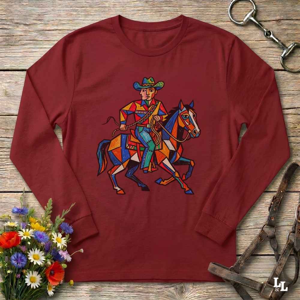 A long-sleeve crewneck t-shirt in maroon red, featuring a colorful geometric graphic of a cowboy riding a horse. The shirt is displayed flat on a rustic wooden background, styled with equestrian leather tack and a small bouquet of wildflowers.