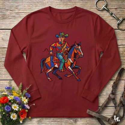 A long-sleeve crewneck t-shirt in maroon red, featuring a colorful geometric graphic of a cowboy riding a horse. The shirt is displayed flat on a rustic wooden background, styled with equestrian leather tack and a small bouquet of wildflowers.