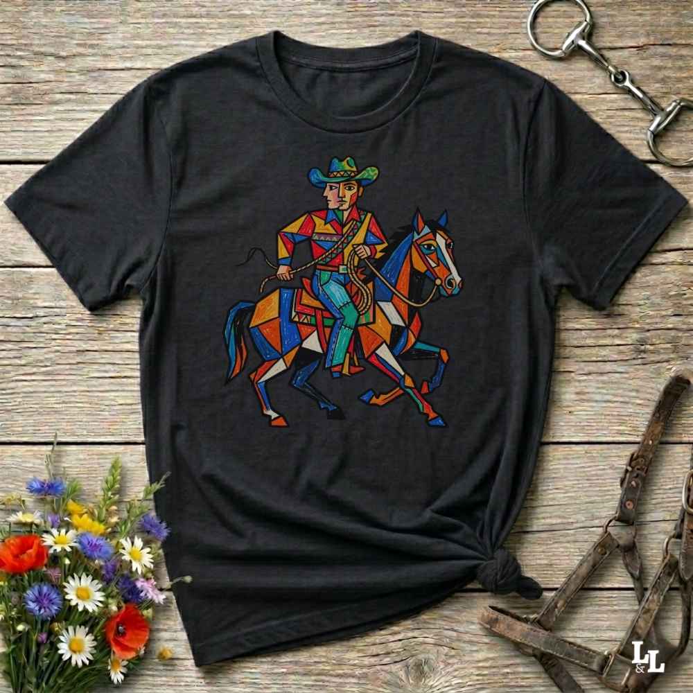 A black short-sleeve graphic t-shirt featuring a colorful geometric illustration of a cowboy riding a horse, laid flat on a rustic wooden surface with decorative flowers and leather tack around it.