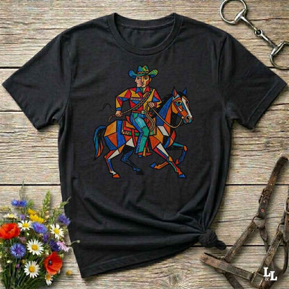 A black short-sleeve graphic t-shirt featuring a colorful geometric illustration of a cowboy riding a horse, laid flat on a rustic wooden surface with decorative flowers and leather tack around it.