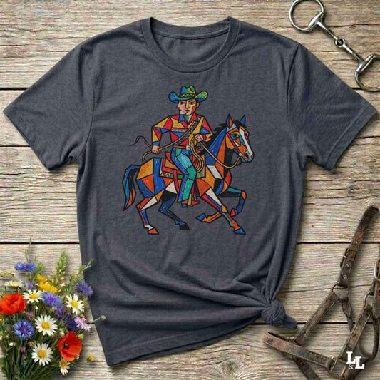 A dark gray heather t-shirt with a colorful graphic of a cowboy riding a horse, styled in a flat-lay on a rustic wooden background with flowers and horse tack.