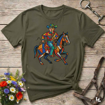 An olive green short-sleeve t-shirt with a colorful geometric graphic of a cowboy riding a horse, displayed on a rustic wooden background with decorative floral and leather props.