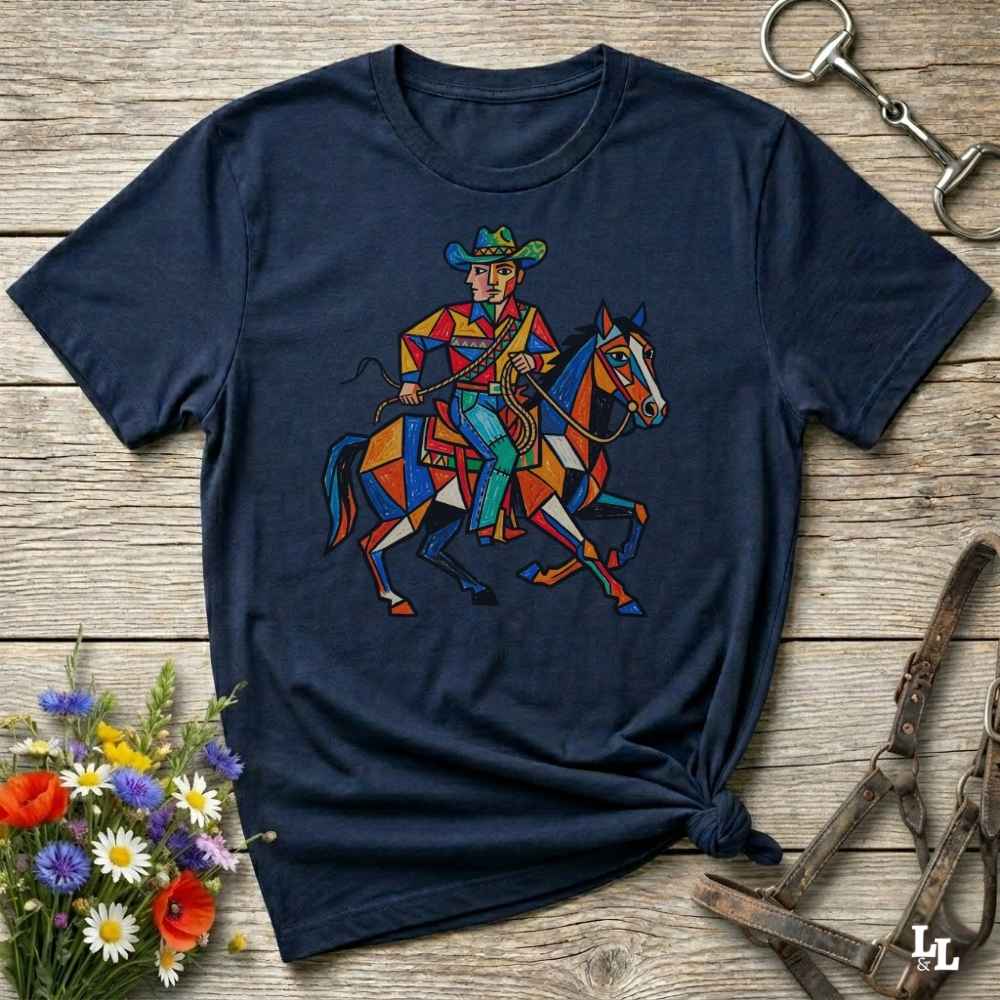 A navy blue short-sleeve t-shirt with a colorful graphic of a cowboy riding a horse, styled on a rustic wooden background with flowers and horse tack.