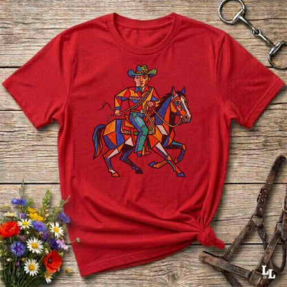 A red short-sleeve t-shirt with a colorful geometric cowboy on a horse graphic, styled flat on a rustic wooden background with a knot at the bottom and equestrian-themed props.