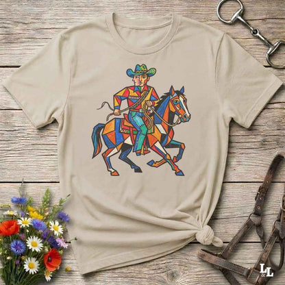 A beige short-sleeve t-shirt with a colorful geometric graphic of a cowboy riding a horse, styled flat on a rustic wooden surface with flowers and leather tack props.