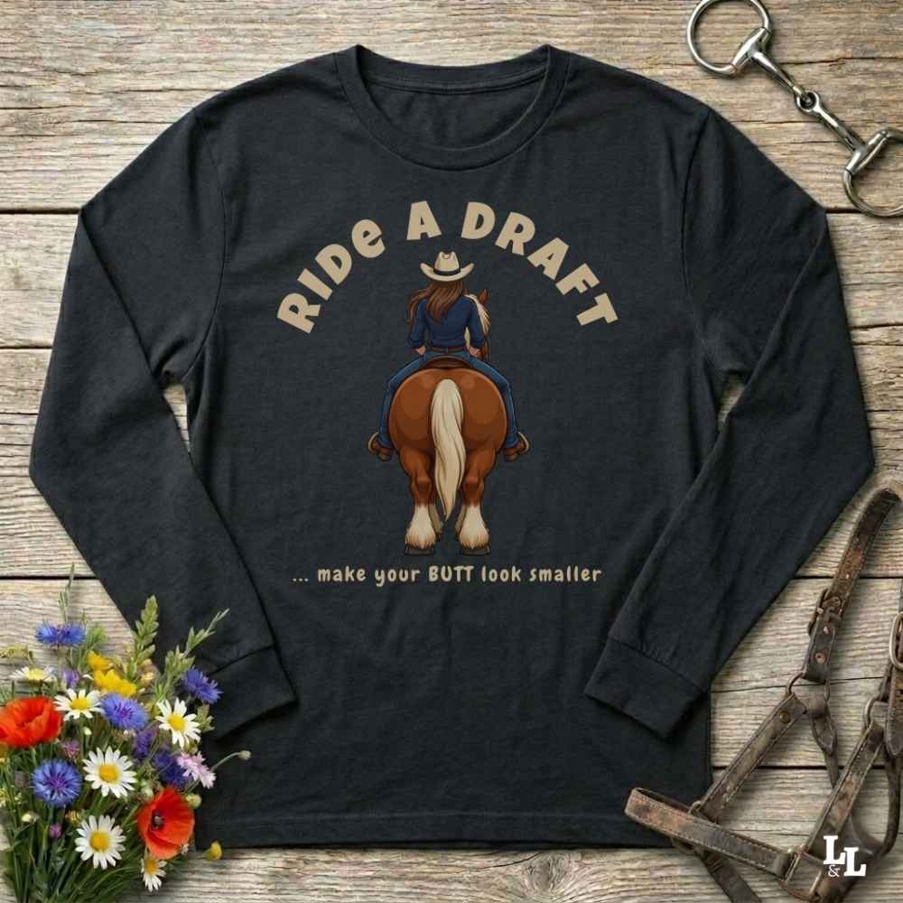 A black long-sleeve graphic t-shirt laid flat on a rustic wooden surface, featuring a cartoon illustration of a cowboy riding a brown horse with the text 'RIDE A DRAFT .