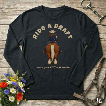 A black long-sleeve graphic t-shirt laid flat on a rustic wooden surface, featuring a cartoon illustration of a cowboy riding a brown horse with the text 'RIDE A DRAFT .