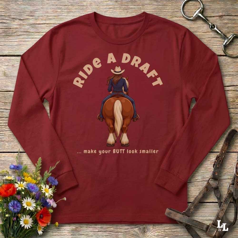 A long-sleeve crewneck t-shirt in maroon red, laid flat on a rustic wooden background. The shirt features a graphic of a cowboy riding a brown horse with the text 'RIDE A DRAFT' and '.
