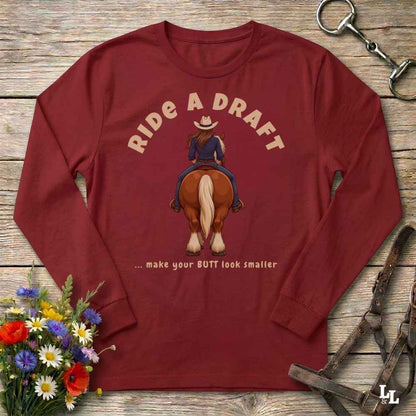 A long-sleeve crewneck t-shirt in maroon red, laid flat on a rustic wooden background. The shirt features a graphic of a cowboy riding a brown horse with the text 'RIDE A DRAFT' and '.