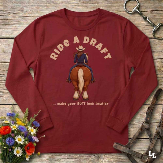 A long-sleeve crewneck t-shirt in maroon red, laid flat on a rustic wooden background. The shirt features a graphic of a cowboy riding a brown horse with the text 'RIDE A DRAFT' and '.