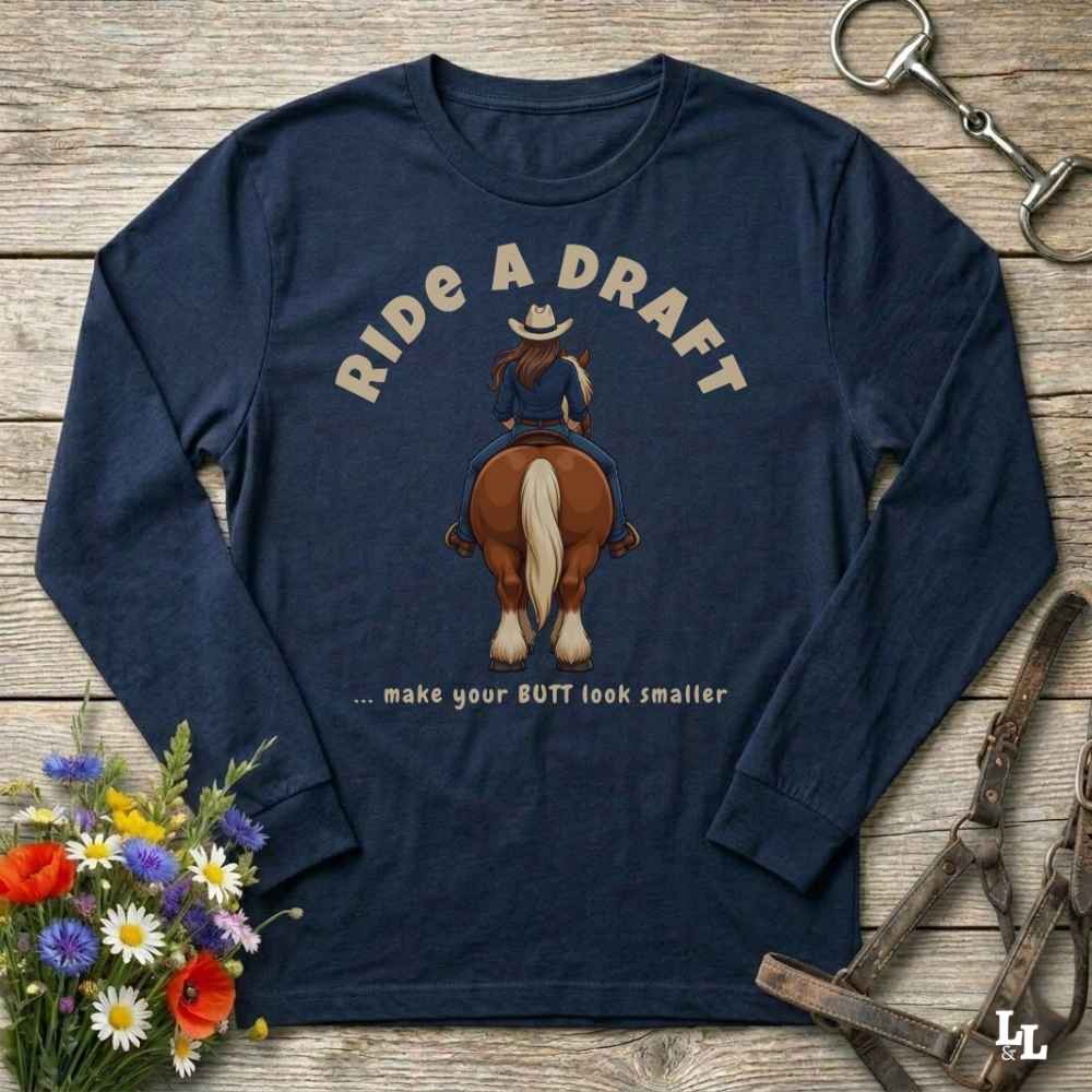 A navy blue long-sleeve graphic t-shirt laid flat on a rustic wooden background, featuring a cowboy riding a horse with the text 'RIDE A DRAFT .