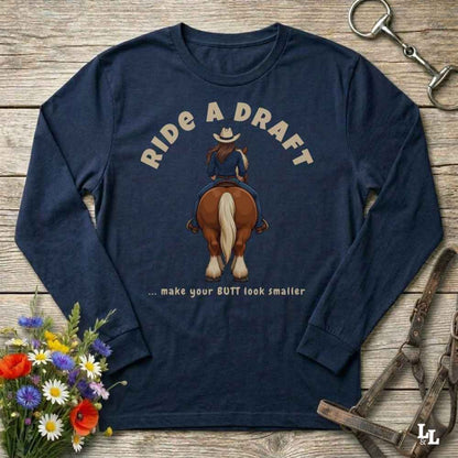 A navy blue long-sleeve graphic t-shirt laid flat on a rustic wooden background, featuring a cowboy riding a horse with the text 'RIDE A DRAFT .
