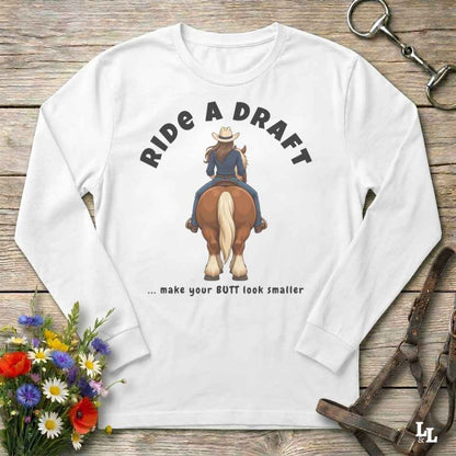 A white long-sleeve t-shirt with a western-themed graphic print, laid flat on a rustic wooden background with equestrian accessories and flowers.