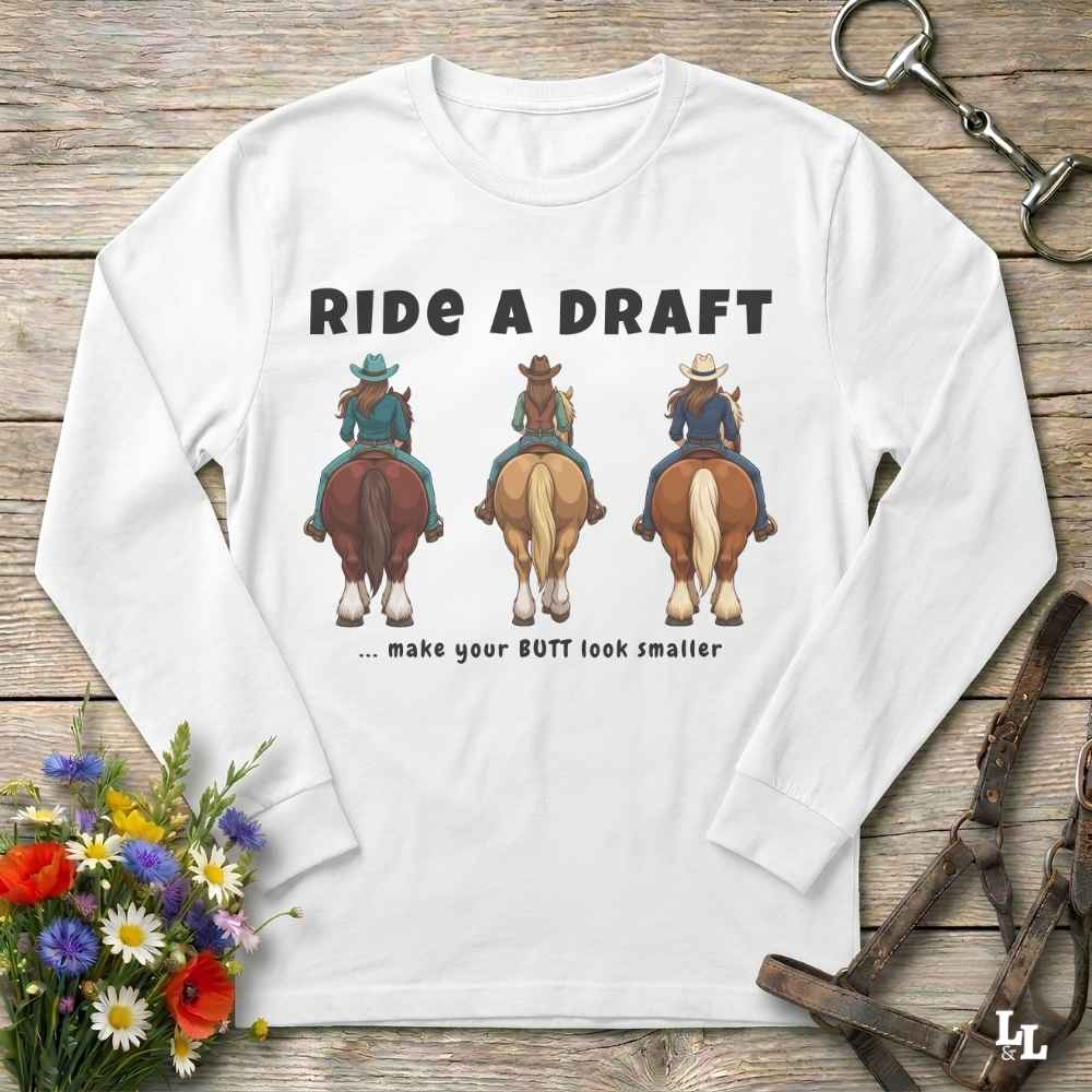 A white long-sleeve t-shirt with a humorous western graphic print, laid flat on a rustic wooden background with equestrian props.