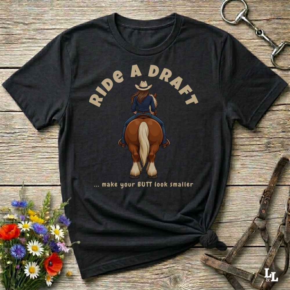 A black graphic t-shirt laid flat on a rustic wooden surface, styled with a small bouquet of wildflowers and equestrian accessories. The shirt features a graphic of a woman riding a brown horse from behind, with the text 'RIDE A DRAFT .