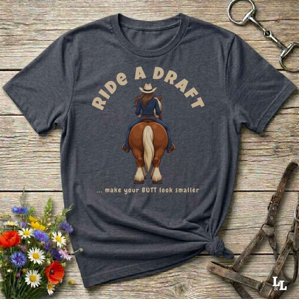 A dark heather gray graphic t-shirt laid flat on a rustic wooden surface. The shirt features a western-themed graphic of a woman riding a brown horse, with the text 'RIDE A DRAFT .