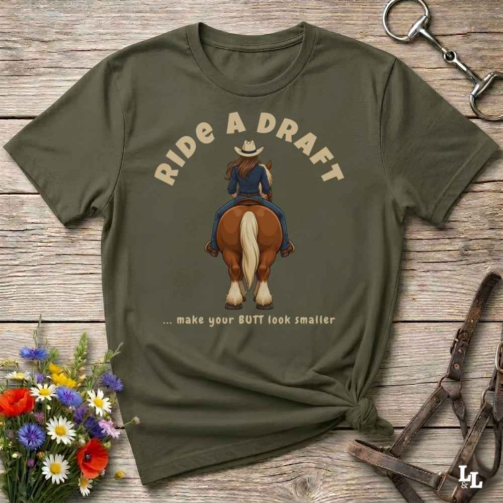 An olive green graphic t-shirt laid flat on a rustic wooden surface, styled with a small bouquet of wildflowers, a horse bit, and a leather bridle. The t-shirt features a graphic of a woman riding a brown horse from behind, with the text 'RIDE A DRAFT .