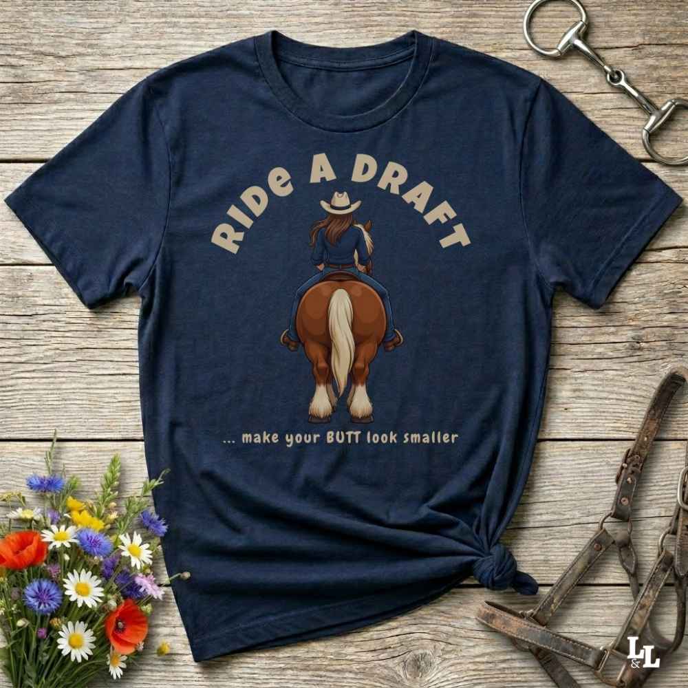 A navy blue graphic t-shirt laid flat on a rustic wooden surface, styled with a small bouquet of wildflowers, a horse bit, and a leather halter. The shirt features a graphic of a woman riding a brown horse from behind, with the text 'RIDE A DRAFT .