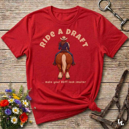 A red short-sleeve graphic T-shirt laid flat on a rustic wooden surface, with a printed illustration of a woman riding a horse from the back and the text 'RIDE A DRAFT .